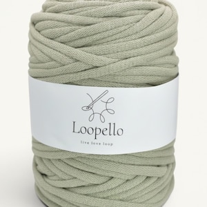 Puede incluir: Una bobina grande de hilo de algodón color verde salvia enrollado en una hebra gruesa y continua. Una etiqueta de papel blanco envuelve el centro, con un dibujo lineal negro de una aguja e hilo con el texto Loopello y live love loop.