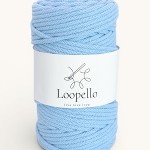 Op de afbeelding: Een grote spoel lichtblauw garen met een wit etiket. Het etiket toont een naald- en draadontwerp, het woord "Loopello" en de zin "live love loop". Het garen is strak opgerold en ziet er zacht uit.