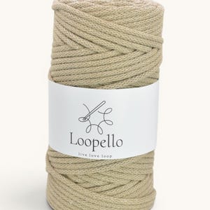 May include: A cylindrical spool of beige macrame cord wrapped in a tight, braided pattern. A white paper label is wrapped around the center, featuring a black line-art icon of a needle and thread, the text Loopello, and the text live love loop.