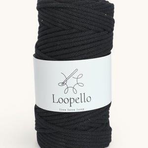 May include: A roll of black macrame cord with a white label wrapped around it. The label features the brand name "Loopello" and the tagline "live love loop" in a simple, elegant font. The cord is tightly wound, showing its texture.