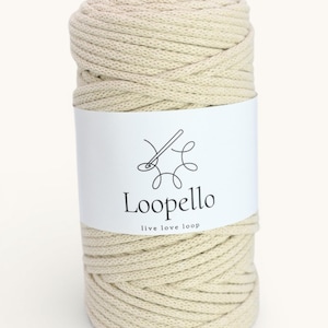May include: A cylindrical spool of off-white macrame cord. A white label wraps around the cord, featuring a logo and the text "Loopello live love loop". The cord is tightly wound, showing a textured surface.