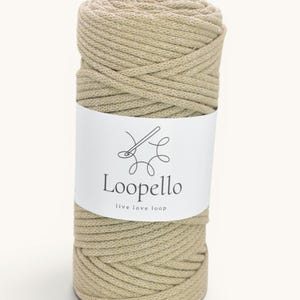 Puede incluir: Una bobina cilíndrica de hilo de cordón beige envuelta en una etiqueta de papel blanco. La etiqueta presenta un dibujo lineal negro de un ganchillo y el texto Loopello, seguido del texto más pequeño live love loop en una fuente negra.