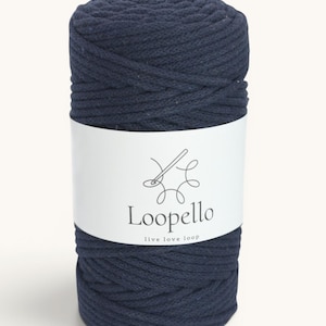 Op de afbeelding: Een cilindrische spoel van marineblauw macramékoord. Een wit etiket wikkelt zich om het koord en toont de merknaam "Loopello" en de slogan "live love loop" in zwarte tekst. Het koord is strak gewikkeld en heeft een getextureerd oppervlak.