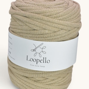 Puede incluir: Una madeja cilíndrica de hilo de algodón de color beige claro envuelta en una banda de papel blanco. La banda presenta el texto negro Loopello, el eslogan live love loop y un gráfico de línea de una aguja e hilo formando un bucle.