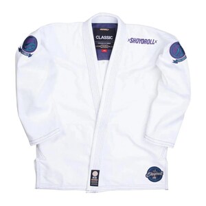 May include: White martial arts uniform with blue stitching and accents. The uniform features the text "CLASSIC" and "SHOYOROLL" in purple. Patches with circular designs are on the sleeves. The uniform is laid flat on a white background.