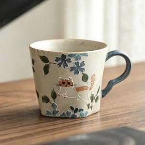 May include: A ceramic mug with a beige background, decorated with blue floral patterns and a cat illustration. The handle is dark blue. The mug sits on a wooden table, with a book and pastries in the background.