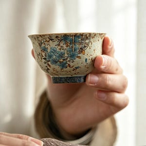 Könnte beinhalten: Eine Keramik-Teetasse mit einem Blumendesign in Blau- und Beigetönen. Die Tasse wird in der Hand einer Person gehalten, wobei die komplizierten Details der gemalten Blumen und die Form der Tasse zur Geltung kommen. Die Tasse hat einen blauen Boden und einen goldenen Rand.