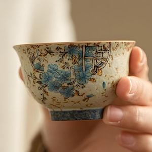 Puede incluir: Una pequeña taza de té de cerámica con un diseño floral en azul y dorado. La taza tiene un fondo moteado blanquecino y una base azul oscuro. La taza está sostenida en la mano de una persona.