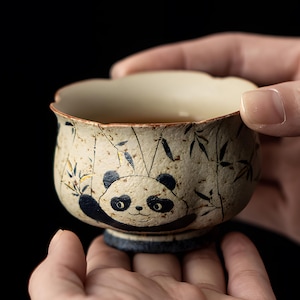 May include: A ceramic tea cup featuring a panda bear design. The cup is beige with a blue and black panda illustration and bamboo leaves. The cup is held in two hands against a black background. The cup has a scalloped rim.