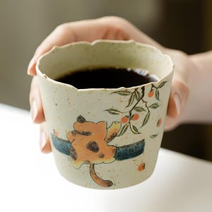 May include: A ceramic cup with a scalloped rim, filled with dark liquid. The cup features a hand-painted design of a cat and branches with orange berries. The cup is held by a person.
