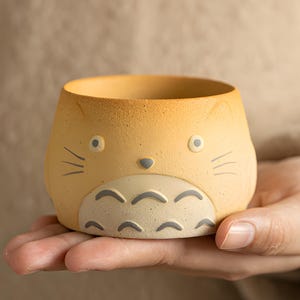 Könnte beinhalten: Ein kleiner, gelber Keramik-Pflanztopf in Form einer Zeichentrickfigur. Der Topf hat einen runden Körper mit einer weiten Öffnung, kleine Augen, eine graue Nase und geschwungene Linien auf der Vorderseite. Der Topf wird in der Hand einer Person gehalten.