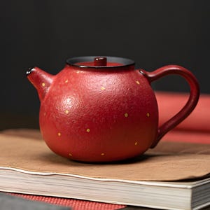 May include: A red ceramic teapot with a rounded body, a curved handle, and a small spout. The teapot is adorned with small, evenly spaced gold dots. The lid is black with a red knob. The teapot is sitting on a book.