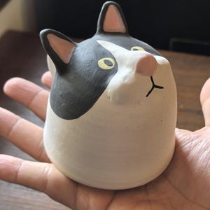 Puede incluir: Una taza de cerámica con forma de gato con un diseño en blanco y negro. La cabeza del gato está inclinada hacia arriba, con ojos amarillos y una nariz rosa. La taza se sostiene en una mano.
