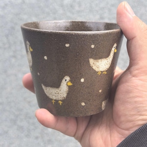 May include: A brown ceramic cup with a speckled texture. The cup is decorated with white ducks and small white dots. The ducks have yellow beaks and feet. The cup is held in a hand.