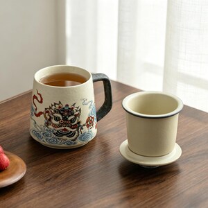 Puede incluir: Una taza de cerámica color crema con un asa negra y una ilustración detallada de una criatura mítica. La taza está llena de té. Un infusor de té a juego se encuentra en un platillo junto a la taza.