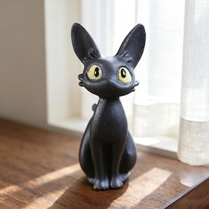 Op de afbeelding: Zwarte kattenbeeldje met grote oren en gele ogen. De kat zit rechtop met zijn poten bij elkaar. Het beeldje is gemaakt van een mat materiaal en staat op een houten oppervlak bij een raam.