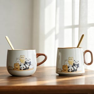 May include: Two white ceramic mugs with brown handles and rims, each featuring a cartoon illustration of bears, pandas, and rabbits. Gold spoons rest inside the mugs. The mugs sit on a wooden surface.