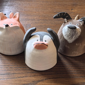 Puede incluir: Tres figuras de animales de cerámica caprichosas: un zorro, un pingüino y una cabra. El zorro es naranja y blanco, el pingüino es negro, blanco y naranja, y la cabra es marrón y blanco. Cada figura tiene un diseño único y caricaturesco.