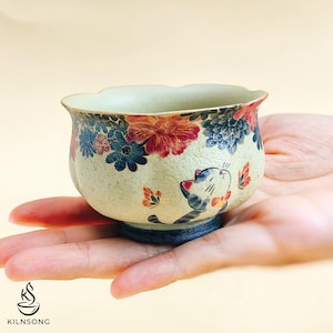 Op de afbeelding: Een kleine, handbeschilderde keramische kom met een bloemen- en kattenontwerp. De kom heeft een crèmekleurige basis met blauwe, rode en oranje bloemen en een grillige kattenillustratie. De kom wordt in een hand gehouden.