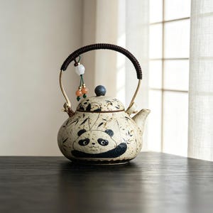 Puede incluir: Una tetera de cerámica con un diseño de oso panda. La tetera tiene un asa marrón y una tapa con un pomo azul oscuro. El panda es blanco y negro, y la tetera tiene un patrón de hojas de bambú. Cuentas decorativas cuelgan del asa.