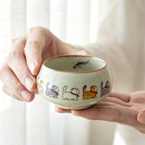以下が含まれることがあります： 淡い緑色の外観の、小さくて丸いセラミックボウル。ボウルの縁には、カラフルな漫画の猫が描かれています。ボウルの中には、小さな魚のイラストが見えます。ボウルは両手で持たれています。