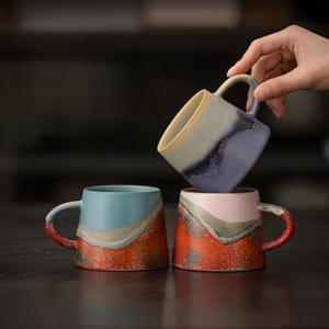 May include: Three ceramic mugs with distinctive colour gradients. One mug is held by a hand. The mugs feature a mix of colours, including blue, red, pink, and cream, with textured surfaces and curved handles. The mugs are designed for beverages.