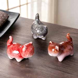 Peut inclure: Trois figurines de chat en céramique rouge, grise et marron. Les chats sont dans une pose ludique, avec leurs queues recourbées. Les figurines sont brillantes et semblent peintes à la main. Elles sont posées sur une surface en bois foncé.