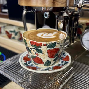 Puede incluir: Una taza y platillo de café de cerámica con un diseño de fresas. La taza está llena de un café con leche, coronado con un diseño de hojas. El juego está sobre una superficie metálica, cerca de una máquina de café.
