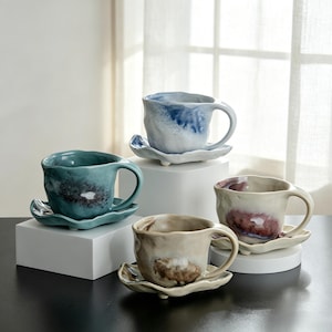 May include: Four handmade ceramic mugs with matching saucers. The mugs are in shades of blue, green, beige, and cream, with organic shapes and textured surfaces. The saucers have a leaf-like design. The mugs are displayed on white pedestals.