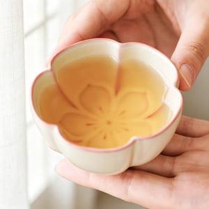 Puede incluir: Una taza de té de cerámica blanca con forma de flor, llena de té. La taza tiene un borde rosa y un diseño floral en el interior. La taza se sostiene con dos manos, con un fondo suave.