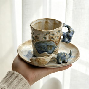 May include: A handmade ceramic coffee cup, saucer, and spoon set. The cup and saucer are beige with blue elephant designs. The cup handle and spoon are shaped like elephants. The set is held in a hand.
