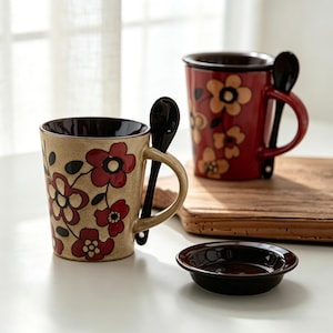 Könnte beinhalten: Zwei Keramikbecher mit Blumendesigns und passenden schwarzen Löffeln. Ein Becher ist beige mit roten Blumen, der andere ist rot mit gelben Blumen. Eine kleine braune Schale befindet sich vor den Bechern. Die Becher haben braune Innenräume und Griffe.