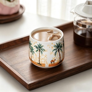 Puede incluir: Una taza de cerámica blanca con una escena desértica con palmeras y un camello. La taza está llena de una bebida marrón claro y cubitos de hielo. La taza está sobre una bandeja de madera.