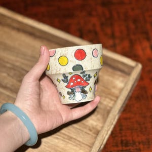 Könnte beinhalten: Ein kleiner, beigefarbener Keramiktopf mit einem skurrilen Pilzdesign. Der Topf hat einen roten Pilz mit weißen Punkten, grünen Blättern und bunten Tupfen. Der Topf wird in einer Hand gehalten, und ein hellblaues Armband ist sichtbar.