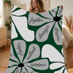 Könnte beinhalten: Eine grün-graue Decke mit floralem Muster und den Wörtern "fighter", "stronger", "courage", "resilient" und "survivor" in Schreibschrift. Die Decke hat einen dunkelgrünen Hintergrund mit großen grauen und weißen Blumenformen.