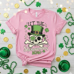 May include: Pink t-shirt with a St. Patrick's Day graphic. The design features a skull wearing a green top hat and an eye patch with a shamrock. The text "LET THE SHENANIGANS BEGIN" is below the skull. Gold coins and green shamrocks are scattered around the shirt.
