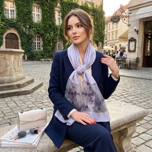 Pode incluir: Uma mulher com um fato azul marinho e um lenço lilás claro com bordados florais. Uma bolsa bege, óculos de sol e revistas estão em um banco de pedra. O fundo apresenta um edifício coberto de hera e uma rua europeia.