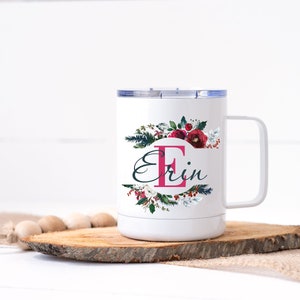 Op de afbeelding: Witte reismok met een bloemenontwerp en de naam "Erin". Het ontwerp bevat een grote roze letter "E" en bordeauxrode bloemen. De mok heeft een wit handvat en een zilveren deksel. Hij staat op een houten schijf.