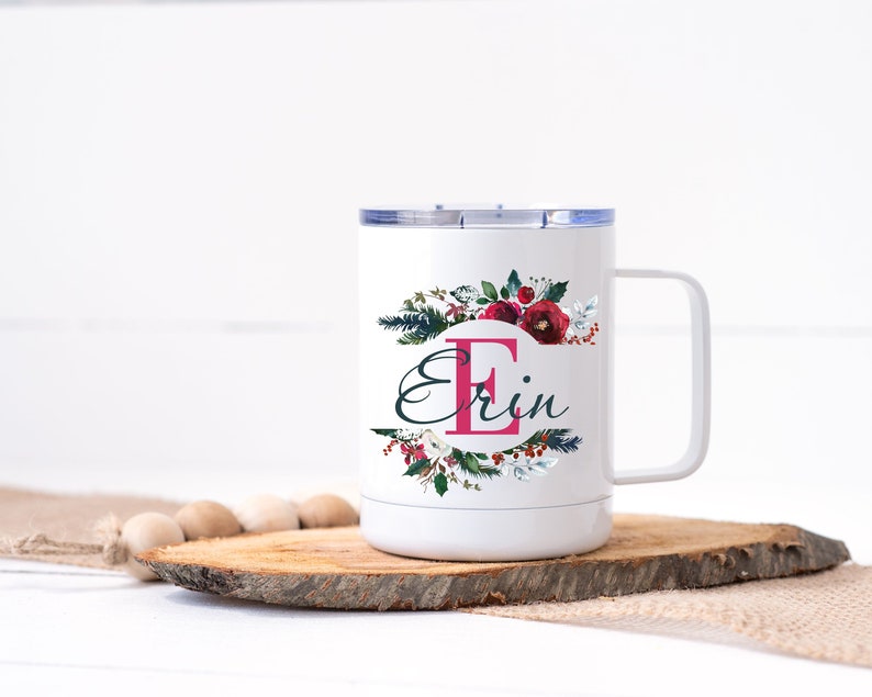 Op de afbeelding: Witte reismok met een bloemenontwerp en de naam "Erin". Het ontwerp bevat een grote roze letter "E" en bordeauxrode bloemen. De mok heeft een wit handvat en een zilveren deksel. Hij staat op een houten schijf.