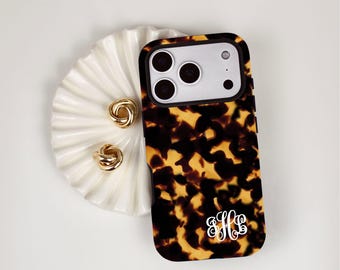 May include: A rectangular smartphone case featuring a brown and black tortoiseshell pattern with a white script monogram of the letters H, L, and B at the bottom. The case has a black border and a cutout for a triple-lens camera system.