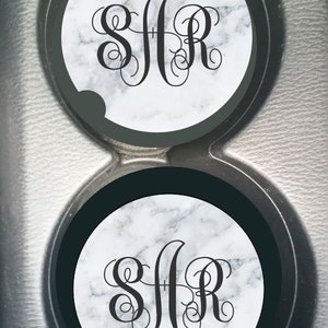 May include: Two round car coasters with a black border and a white marble design. The coasters feature a black monogram with the letters "SJR" in a cursive font.