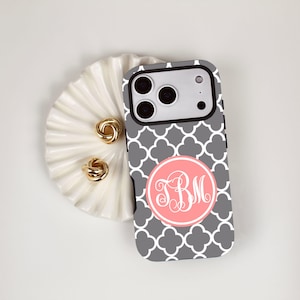 May include: A smartphone case featuring a gray and white quatrefoil pattern with a central coral pink circular emblem containing the white script monogram letters TBM. The case has a black camera cutout area with three circular lenses.