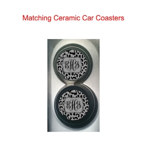 May include: Two black and white leopard print ceramic car coasters with a monogram in a gray square in the center. The coasters are in a car cup holder.