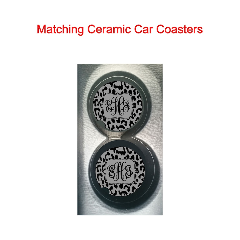 May include: Two black and white leopard print ceramic car coasters with a monogram in a gray square in the center. The coasters are in a car cup holder.