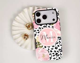May include: A smartphone case featuring a black and white leopard print pattern, accented with soft pink floral illustrations. A central circular emblem displays the letter M and the name Monica in a black script font against a light pink background.