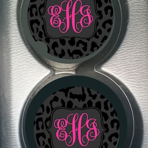 May include: Two black and gray leopard print car coasters with a pink monogram "LLS" in the center of each coaster.