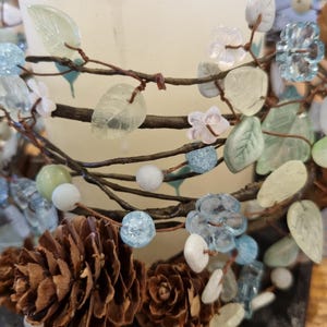 May include: A candle holder featuring a white candle, decorated with faux branches, pinecones, and glass beads in blue, green, and white. The design incorporates natural elements, creating a decorative piece for the home.