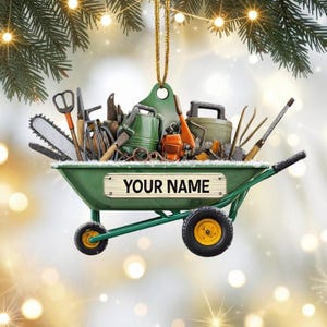May include: A green wheelbarrow ornament filled with gardening tools, including a saw, watering can, and clippers. The ornament has a gold hanging string and the text "YOUR NAME" on the front. The wheels are black with yellow centers.