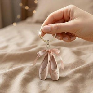May include: A ballet-themed keychain featuring two miniature pink ballet slippers with satin ribbons. The slippers have the name "Olivia" and the year "2025" printed on them. The keychain is attached to a silver ring.