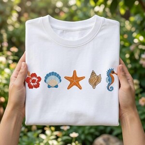 May include: White t-shirt with embroidered beach-themed designs. Features a red hibiscus flower, a blue and beige seashell, an orange starfish, a brown conch shell, and a blue and orange seahorse. The shirt is held up by two hands.
